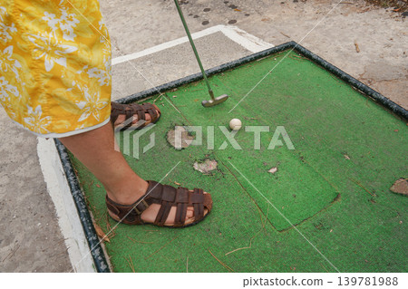 Young man playing minigolf on old, worn hotel resort green field, closeup to his feet in sandals, metal club and ball 139781988