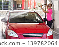 Young woman wearing pink t shirt and sunglasses cleaning her car in self service carwash, spraying water over front windshield and roof 139782064