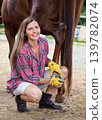 Young woman in shirt crouching as she's cleaning horse hoof with metal horseshoe 139782074