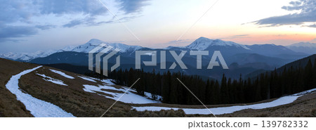 Panorama of beautiful spring morning in Carpathian mountains. Valley with dry grass and snow, transparent fresh air, dense evergreen forest and soft sun glow over distant snow-covered mountain range. Panorama of beautiful spring morning in Carpathian mountains. Valley with dry grass and snow, transparent fresh air, dense evergreen forest and soft sun glow over distant snow-covered mountain range. 139782332