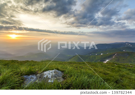 Wide summer mountain view at sunrise. Glowing orange sun raising in blue cloudy sky over green grassy hill with big rock and distant mountain range covered with morning mist. Beauty of nature concept. Wide summer mountain view at sunrise. Glowing orange sun raising in blue cloudy sky over green grassy hill with big rock and distant mountain range covered with morning mist. Beauty of nature concept. 139782344