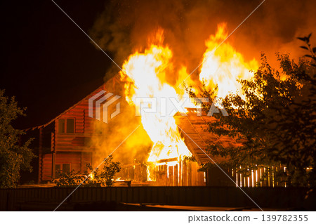 Burning wooden house at night. Bright orange flames and dense smoke from under the tiled roof on dark sky, trees silhouettes and residential neighbor cottage background. Disaster and danger concept. Burning wooden house at night. Bright orange flames and dense smoke from under the tiled roof on dark sky, trees silhouettes and residential neighbor cottage background. Disaster and danger concept. 139782355