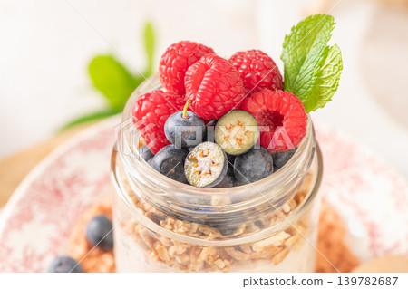 Glass jar filled with creamy overnight oats and chia seeds layered with granola and topped with fresh raspberries and blueberries served with a wooden spoon and mint garnish in a bright rustic kitchen Glass jar filled with creamy overnight oats and chia seeds layered with granola and topped with fresh raspberries and blueberries served with a wooden spoon and mint garnish in a bright rustic kitchen 139782687