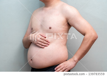 Man with round belly poses with hand on stomach against plain background. Humorous body gesture imitates a pregnancy portrait in a casual indoor setting. Man with round belly poses with hand on stomach against plain background. Humorous body gesture imitates a pregnancy portrait in a casual indoor setting. 139783559