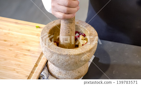 A chief puts chilies and garlics in the stone mortar and pounding with stone pestle on the counter. Preparing food ingredients. A chief puts chilies and garlics in the stone mortar and pounding with stone pestle on the counter. Preparing food ingredients. 139783571