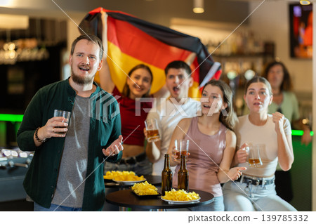 Group of fans of Germany team with a flag are watching the match on TV in beer bar. They are worried and supporting their Group of fans of Germany team with a flag are watching the match on TV in beer bar. They are worried and supporting their 139785332