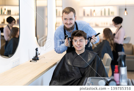 Young man barbershop employee stands next to male client, discussing details of haircut Young man barbershop employee stands next to male client, discussing details of haircut 139785358