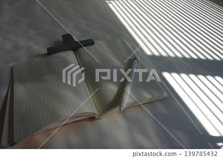 A desk with a grid notebook and pen, where light and shadow fall through the blinds. A desk with a grid notebook and pen, where light and shadow fall through the blinds. 139785432