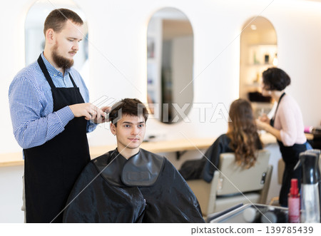 Young man barbershop employee stands next to male client, discussing details of haircut 139785439