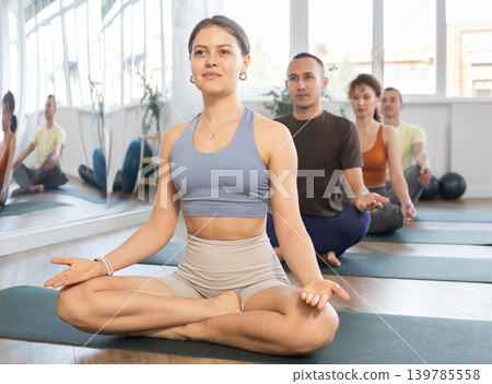 Girl yoga trainer perform doing traditional lotus position with group of students in gym Girl yoga trainer perform doing traditional lotus position with group of students in gym 139785558