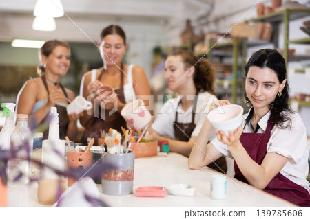 Young woman sits near table and decorates clay ceramic craft vase, plate Young woman sits near table and decorates clay ceramic craft vase, plate 139785606