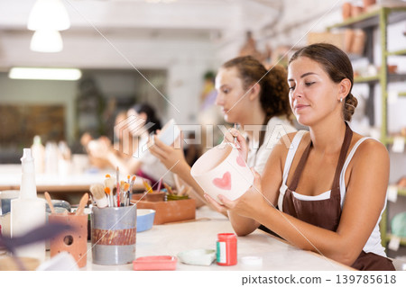 Young woman sits near table and decorates clay ceramic craft vase, plate 139785618