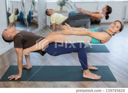 Young men and women maintaining healthy lifestyle practicing partner yoga at group lesson, training balance pose at gym 139785620