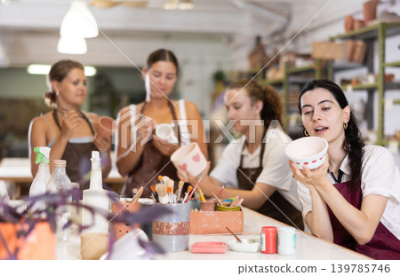 Young woman sits near table and decorates clay ceramic craft vase, plate Young woman sits near table and decorates clay ceramic craft vase, plate 139785746