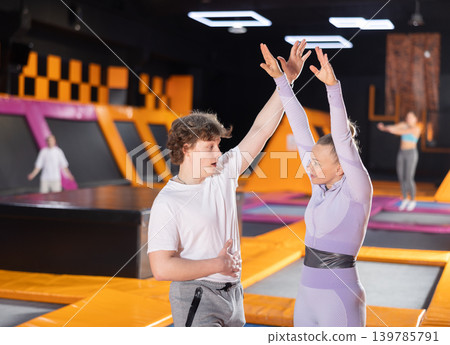 Skilled female trainer helping young guy warming up and preparing for exercising on trampolines in sports center Skilled female trainer helping young guy warming up and preparing for exercising on trampolines in sports center 139785791