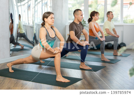 Lovers of active lifestyle on yoga mat perform doing version of warrior pose in gym Lovers of active lifestyle on yoga mat perform doing version of warrior pose in gym 139785974