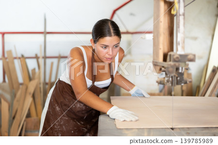 Girl employee of workshop cuts wooden board into parts using saw Girl employee of workshop cuts wooden board into parts using saw 139785989