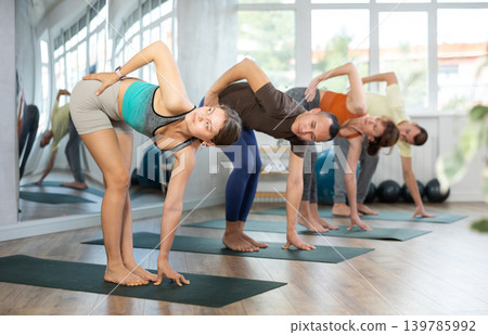 Lovers of active lifestyle on yoga mat perform doing parivritta uttatasana in gym Lovers of active lifestyle on yoga mat perform doing parivritta uttatasana in gym 139785992