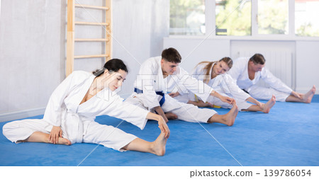 Young woman wearing kimono practicing leg stretching position in sports hall Young woman wearing kimono practicing leg stretching position in sports hall 139786054