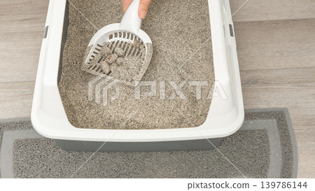 Hand using a scoop to clean a gray cat litter box placed on a floor mat in a well-lit indoor environment 139786144