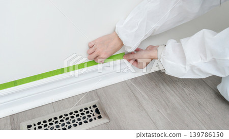 Person applying painter's tape along the baseboard in a home renovation project, with wooden flooring and air vent visible in the background Person applying painter's tape along the baseboard in a home renovation project, with wooden flooring and air vent visible in the background 139786150