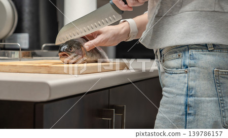 Woman in gray sweater slicing raw fish fillet on wooden cutting board in kitchen. Home cooking preparation, seafood filleting, fresh fish processing, kitchen activity. Woman in gray sweater slicing raw fish fillet on wooden cutting board in kitchen. Home cooking preparation, seafood filleting, fresh fish processing, kitchen activity. 139786157