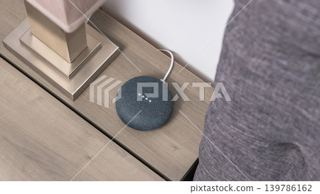 Smart home speaker placed on a wooden table beside a lamp, with a gray fabric surface visible in the background Smart home speaker placed on a wooden table beside a lamp, with a gray fabric surface visible in the background 139786162