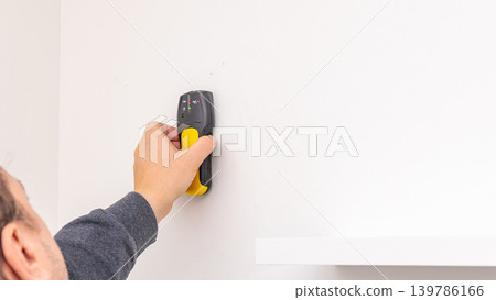 Mans hand pressing yellow stud finder on white wall above installed black shelf bracket. Home improvement, verifying wall support, DIY post-installation check, tool usage. Mans hand pressing yellow stud finder on white wall above installed black shelf bracket. Home improvement, verifying wall support, DIY post-installation check, tool usage. 139786166
