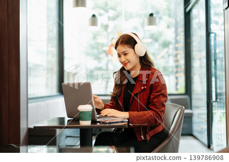 professional Asian woman wears headphones while working on her laptop at a modern cafe, representing online education, remote meetings professional Asian woman wears headphones while working on her laptop at a modern cafe, representing online education, remote meetings 139786908