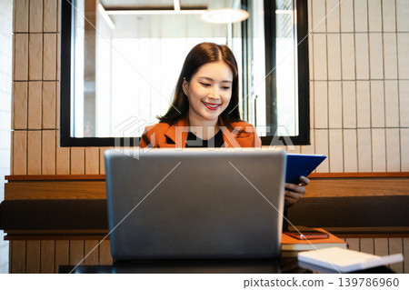 Asian businesswoman writing notes while working on laptop in modern office. Asian businesswoman writing notes while working on laptop in modern office. 139786960