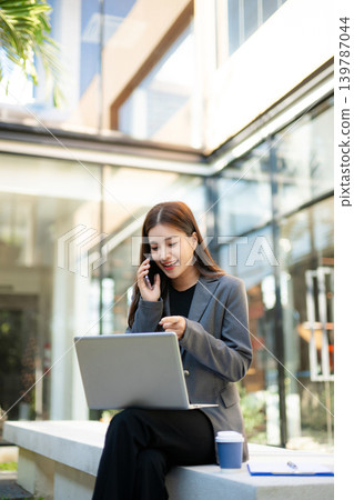 Smiling businesswoman talking on smartphone while using laptop outdoors. Professional corporate lifestyle, remote work, technology, communication and modern business concept. Smiling businesswoman talking on smartphone while using laptop outdoors. Professional corporate lifestyle, remote work, technology, communication and modern business concept. 139787044