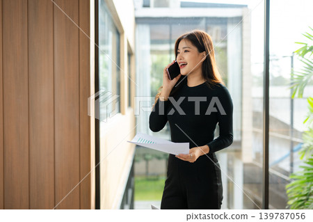 Asian businesswoman using laptop and smartphone while reviewing documents in a modern workspace, symbolizing productivity, remote work Asian businesswoman using laptop and smartphone while reviewing documents in a modern workspace, symbolizing productivity, remote work 139787056
