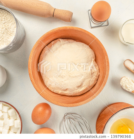 Dough in wooden bowl with baking ingredients and kitchen utensils on light background Dough in wooden bowl with baking ingredients and kitchen utensils on light background 139790368