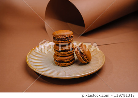Chocolate macarons sit on a round plate against a brown backdrop Chocolate macarons sit on a round plate against a brown backdrop 139791312