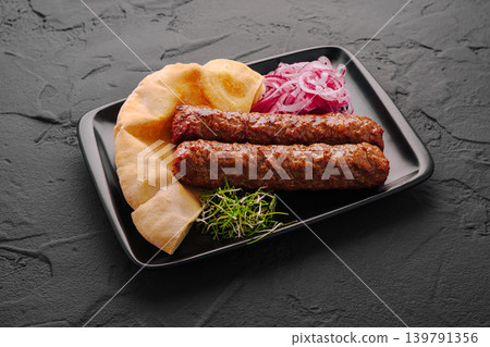 Delicious kebabs served with bread, onions, and greens on a black plate Delicious kebabs served with bread, onions, and greens on a black plate 139791356