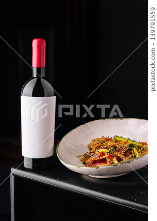 Wine bottle on a table next to a dish of colorful food in a dark setting Wine bottle on a table next to a dish of colorful food in a dark setting 139791559