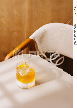 Classic cocktail with orange zest and ice cube on a table near a chair in a cozy indoor setting Classic cocktail with orange zest and ice cube on a table near a chair in a cozy indoor setting 139792379