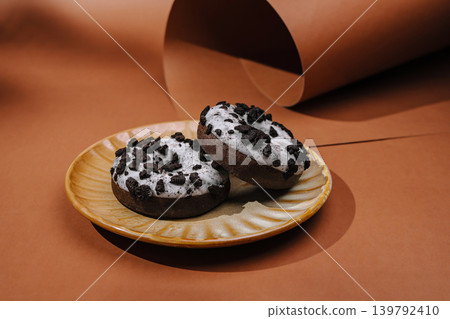 Chocolate donuts with white icing and cookie crumbles on a plate with black and brown background in simple setting Chocolate donuts with white icing and cookie crumbles on a plate with black and brown background in simple setting 139792410