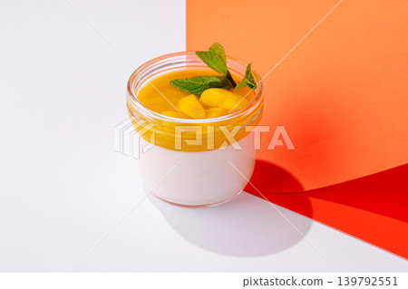 Mango dessert in a glass jar sits on a white surface beside colorful paper Mango dessert in a glass jar sits on a white surface beside colorful paper 139792551
