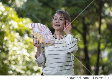 A senior woman cools herself off by fanning herself with a fan in a park surrounded by fresh greenery. A senior woman cools herself off by fanning herself with a fan in a park surrounded by fresh greenery. 139793094