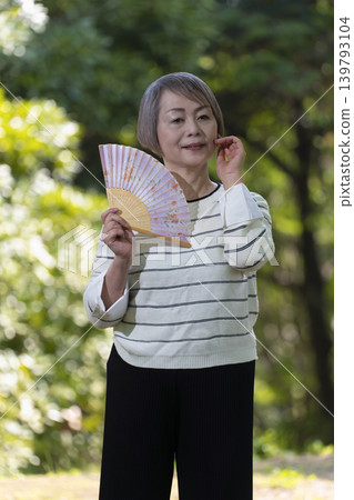 一位老婦人在綠樹環繞的公園裡用扇子搧風降溫。 一位老婦人在綠樹環繞的公園裡用扇子搧風降溫。 139793104