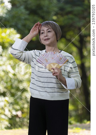 一位老婦人在綠樹環繞的公園裡用扇子搧風降溫。 一位老婦人在綠樹環繞的公園裡用扇子搧風降溫。 139793105