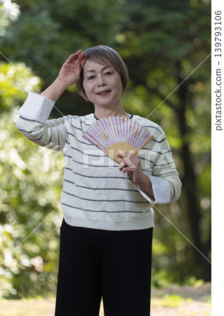 一位老婦人在綠樹環繞的公園裡用扇子搧風降溫。 一位老婦人在綠樹環繞的公園裡用扇子搧風降溫。 139793106