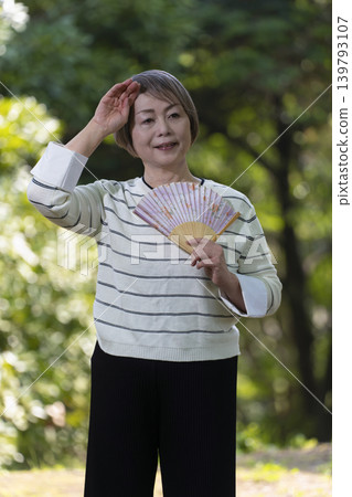 A senior woman cools herself off by fanning herself with a fan in a park surrounded by fresh greenery. A senior woman cools herself off by fanning herself with a fan in a park surrounded by fresh greenery. 139793107