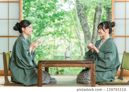 一位女士和她的朋友穿著浴衣,在一家傳統日式旅館的日式房間裡喝清酒。 一位女士和她的朋友穿著浴衣,在一家傳統日式旅館的日式房間裡喝清酒。 139793318
