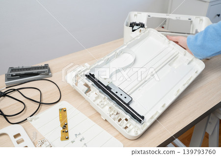 Hands working on a disassembled flatbed scanner laid out on a light wooden desk, exposing internal parts, ribbon cable and circuit board during maintenance or DIY electronics repair in a home office. Hands working on a disassembled flatbed scanner laid out on a light wooden desk, exposing internal parts, ribbon cable and circuit board during maintenance or DIY electronics repair in a home office. 139793760