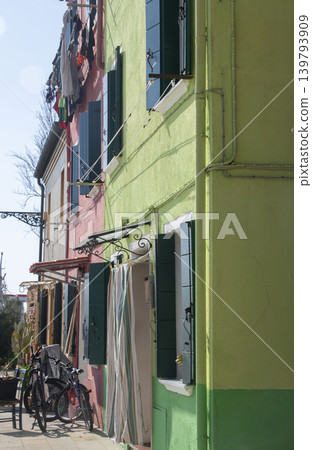 A lively scene of people in front of their homes on Burano Island, with colorful houses lined up and bicycles and other items in the entranceway. A lively scene of people in front of their homes on Burano Island, with colorful houses lined up and bicycles and other items in the entranceway. 139793909