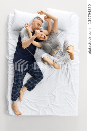 Laughing couple moving on white bed from overhead view while woman holds tablet with feet. 139794820