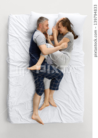 Happy couple lying on white bed from overhead view and laughing together. Happy couple lying on white bed from overhead view and laughing together. 139794833