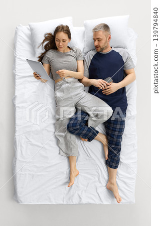 Couple lying on white bed from overhead view using tablet and smartphone. Couple lying on white bed from overhead view using tablet and smartphone. 139794840